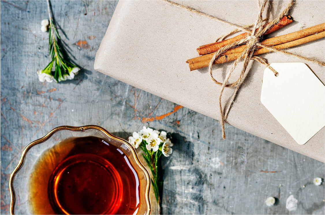 Wrapped gift with cinnamon sticks and white flowers beside a golden saucer of amber tea